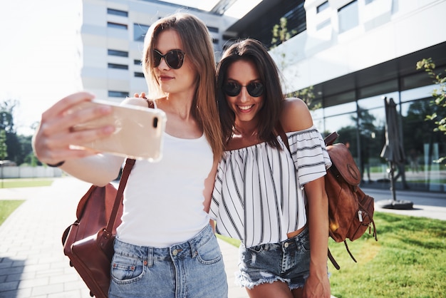 Two beautiful happy young student girl with backpack near the campus of university and make selfie photo. Education and leisure concept