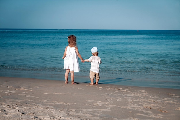 Two beautiful little children walk along the seashore in a day timer and hold their hands. They are in white clothes and hat