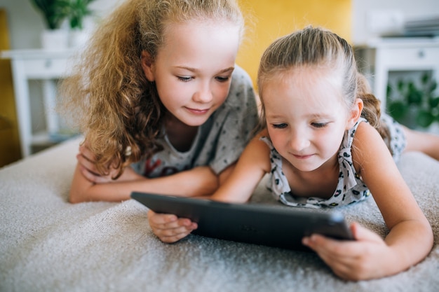 Two beautiful little sisters lying in the bed and look at the screen of a tablet, smart kids using smart technology