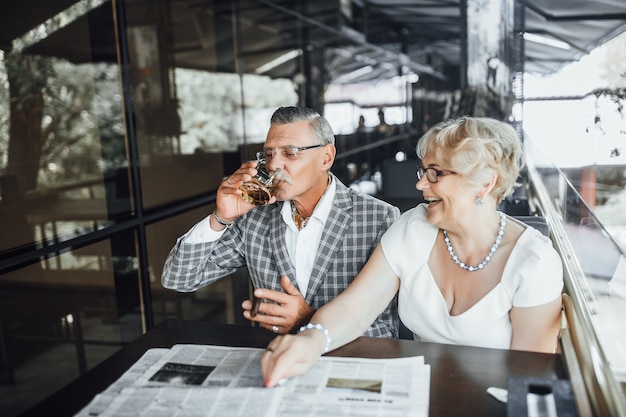 Two beautiful seniors couple drinking wine at summer terrace ,and reading newspaper at this time