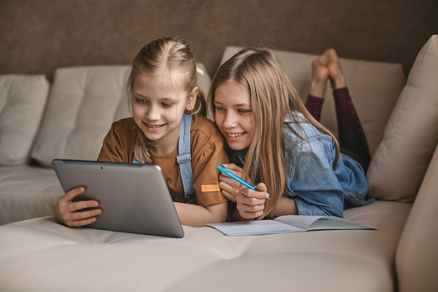 Two beautiful sisters do their homework during quarantine. Children use gadgets for learning. 