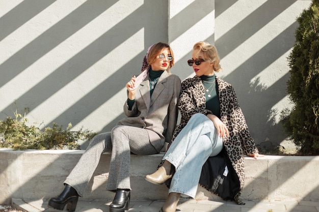 Two beautiful stylish girlfriends of the model in fashionable clothes with leopard coats clush jeans a suit and shoes sit in the city with sunlight Urban vintage style and fashion outfit