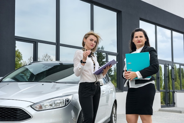 Two beautiful women handshaking near new car standing outside and smilling