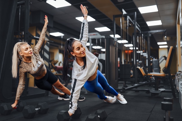 Two beautiful women with athletic figures perform a side plank pose in gym