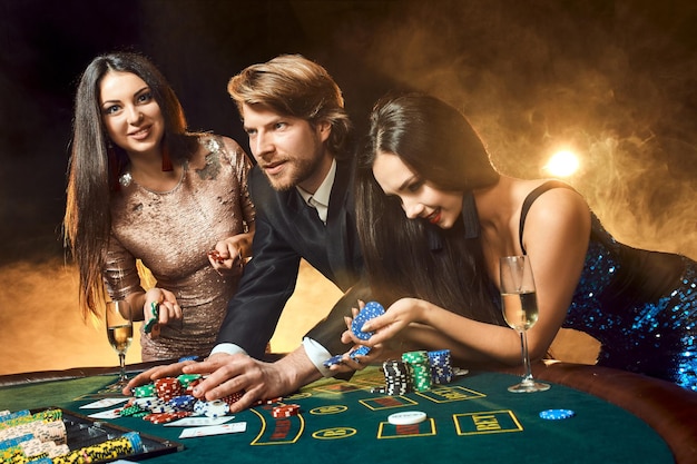 Two beautiful women and young man play on poker table in casino, focus on man and brunette