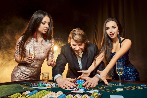 Two beautiful women and young man play on poker table in casino, focus on man and brunette