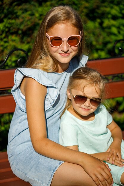 Two beautiful young girls in sun-protected glasses sit on a park bench