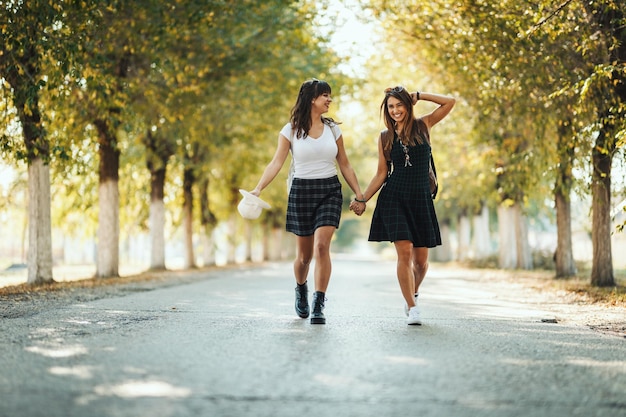 Two beautiful young smiling women with backpacks on their back are walking along the autumn sunny avenue holding hands.