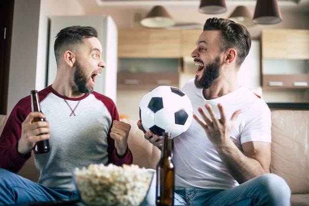 Two best friends and fans of football watching some sport match on the TV and drinking beers and eating snacks while cheering for the team on the couch