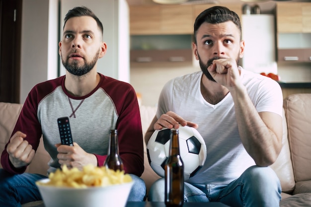 Two best friends and fans of football watching some sport match on the TV and drinking beers and eating snacks while cheering for the team on the couch