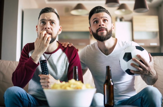 Two best friends and fans of football watching some sport match on the TV and drinking beers and eating snacks while cheering for the team on the couch