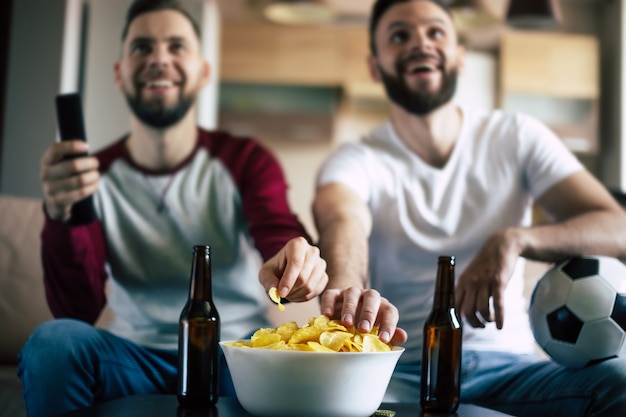 Two best friends and fans of football watching some sport match on the TV and drinking beers and eating snacks while cheering for the team on the couch