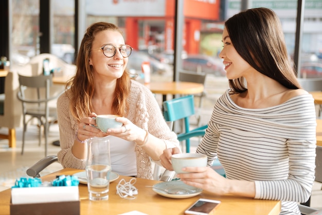 Two besties. Couple of girlfriends are looking at each other while sitting at cafe and holding cups with coffee.