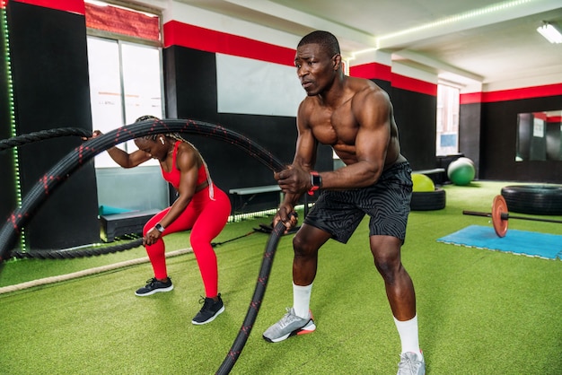 Two black bodybuilder athletes in a sports gym doing an aerobic rope exercise with a lot of effort to strengthen arms and chest. Concept of strengthening the body in gym