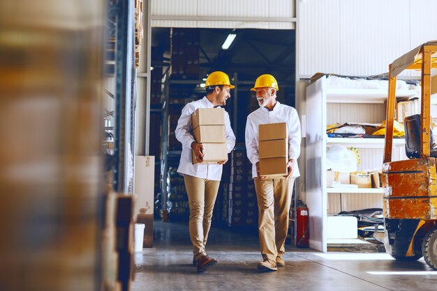 Two blue collar workers in white uniforms and with yellow helmets on heads relocating heavy boxes in warehouse.