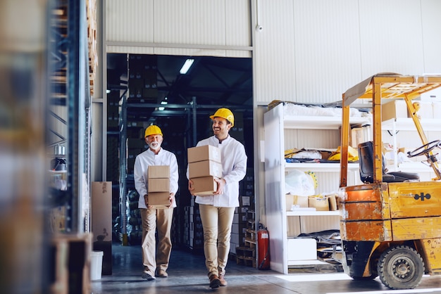 Two blue collar workers in white uniforms and with yellow helmets on heads relocating heavy boxes in warehouse.