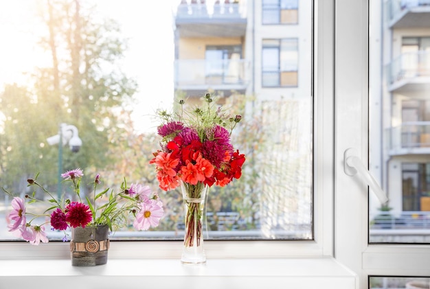 Two bouquets of autumn flowers asters begonia cosmos geranium thistle on a white windowsill against the background of the autumn cityscape