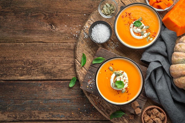 Two bowls of pumpkin cream soup with basil cream and pumpkin seeds on a brown background