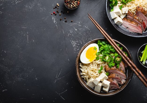 Two bowls of tasty Asian noodle soup ramen with broth, tofu, pork, egg on black rustic stone background, space for text, close up, top view. Hot tasty Japanese ramen soup for dinner with copy space