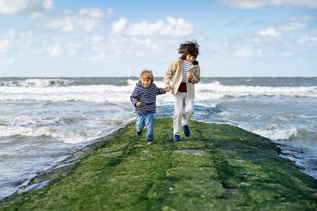 Two brothers holding hands are running on a breakwater at the North Sea. Laughing boys spending weekend at the  seaside in Belgium, Knokke. Siblings friendship.