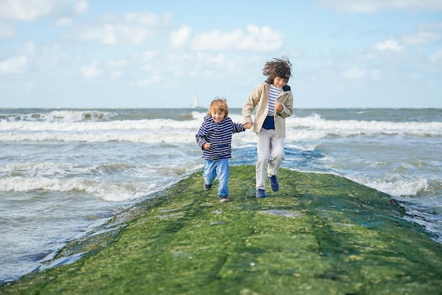 Two brothers holding hands are running on a breakwater at the North Sea Laughing boys spending weekend at the seaside in Belgium Knokke Siblings friendship