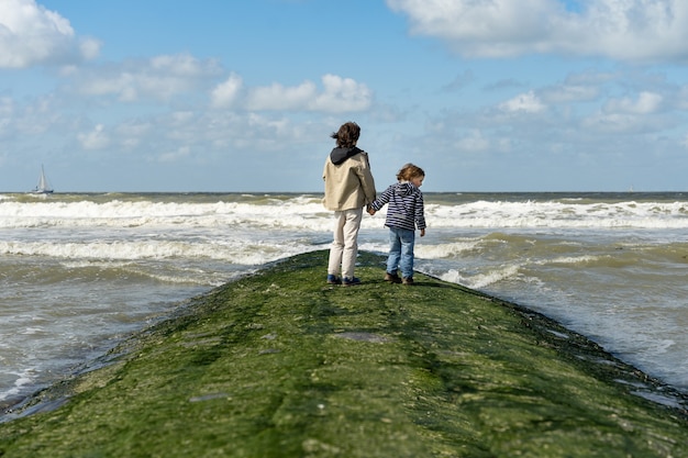 Two brothers holding hands are staying on a breakwater at the North Sea. Boys spending weekend at the  seaside in Belgium, Knokke. Siblings friendship.