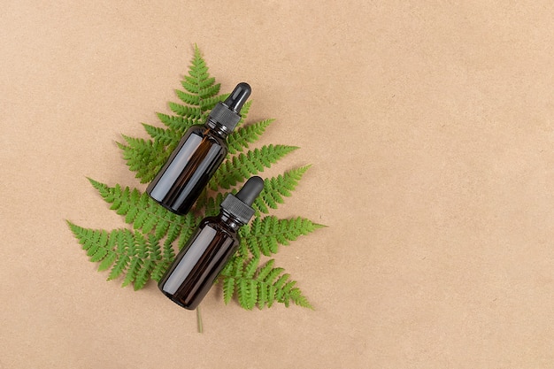 Two brown glass bottles with serum, essential oil or other cosmetic product and green fern leaves on craft beige background. Natural Organic Spa Cosmetic Beauty concept Top view Copy space.