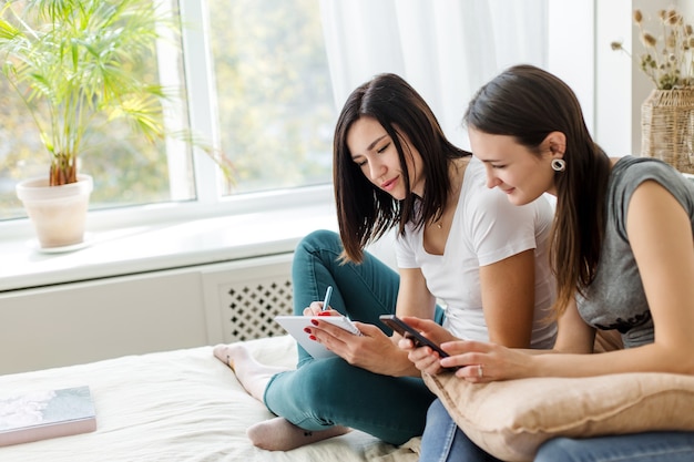 Two brunette girls are sitting talking, holding a book and a phone. 