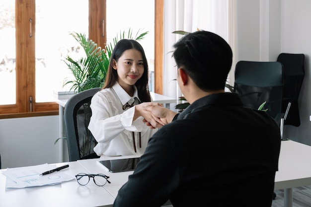 Two business people handshake at meeting table in office together with confident Young asian businessman and businesswoman workers express agreement of investment deal