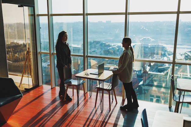 Two business women are standing against the background of a large window in a modern office with a panoramic view of the city Modern business women work remotely