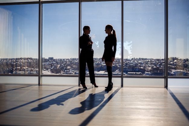 Two business women enjoying the city view and talking while standing by the large window in the office