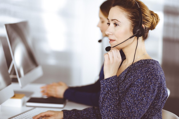 Two busineswomen have conversations with the clients by headsets, while sitting at the desk in a modern office. Diverse people group in a call center. Telemarketing and customer service.