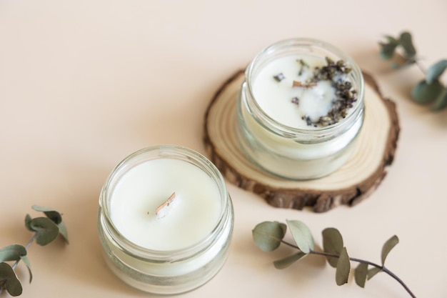 Two candmade candles from paraffin and soy wax in glass with wooden wick and dry herbal isolated on pastel beige background Flat lay top view copy space