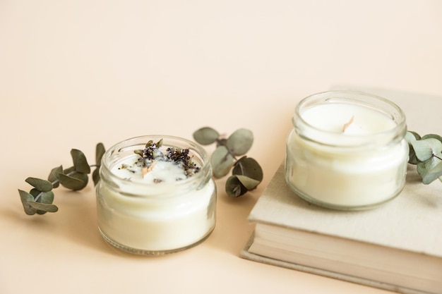 Two candmade candles from paraffin and soy wax in glass with wooden wick and dry herbal isolated on pastel beige background Flat lay top view copy space