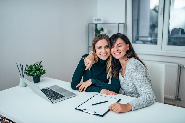 Two casual business women hugging while sitting at work. Friends working together.