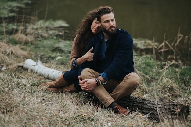 Two caucasian lovers are sitting on the shore of the lake. Young couple is hugging on autumn day outdoors.  Valentine's Day.