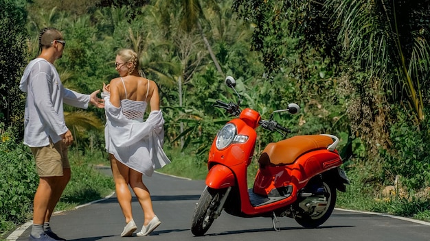 Two caucasian tourist woman man dance near scooter. Love couple on red motorbike in white clothes, sunglasses on forest road trail trip. Dancing road. Motorcycle rent. Asia Thailand ride tourism.