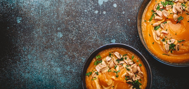 Two ceramic bowls with pumpkin cream soup decorated with peanuts on rustic stone background with spoon from above Autumn cozy dinner concept xD