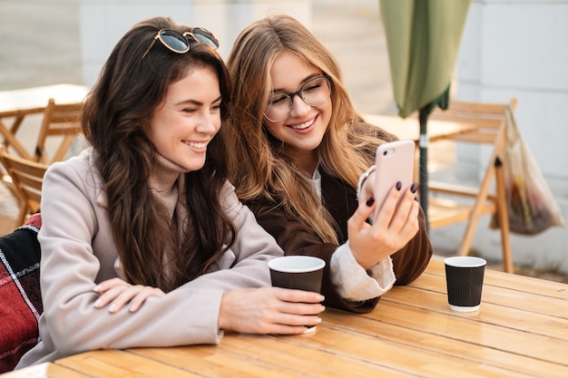 Two cheerful attractive women friends sitting at the cafe outdoors, looking at mobile phone while drinking coffee