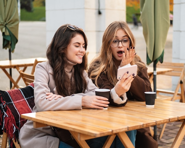 Two cheerful attractive women friends sitting at the cafe outdoors, looking at mobile phone while drinking coffee