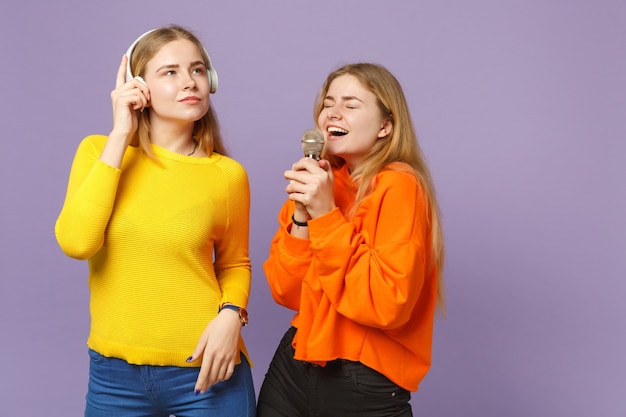 Two cheerful blonde twins sisters girls in vivid clothes listen music with headphones, sing song in microphone isolated on violet blue wall. People family lifestyle concept. 