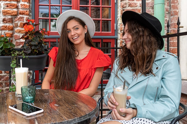 Two cheerful charming young women talking and drinking coffee in outdoor cafe