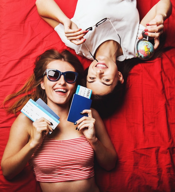 Two cheerful friends girls holding passports plane tickets and globe lying on red bed indoor travel concept