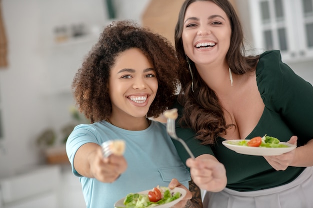 Two cheerful girlfriends offering to try vegetable salad