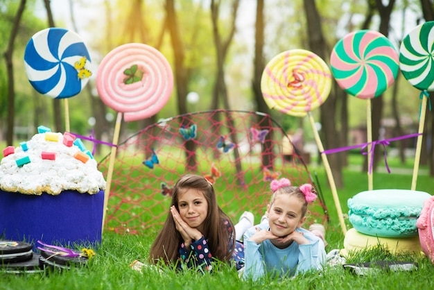 Two cheerful girls lie on the green grass and smile in the scenery from huge sweets of lollipops and cakes