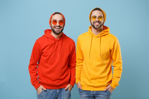 Two cheerful hipster men guys in fashion red yellow clothes eyeglasses posing isolated on pastel blue wall  portrait. People emotions lifestyle concept. 