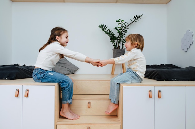 Two cheerful siblings sitting on wooden elevation in front of each other holding hands smiling Side view