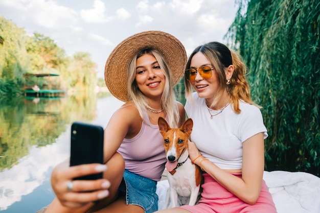 Two cheerful woman friends resting outdoor on lake pier with dog, making photo, selfie