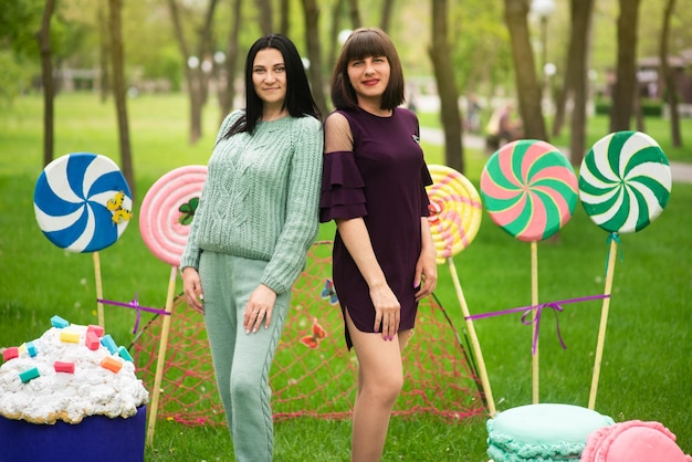 Two cheerful women standing on the green grass in the scenery from huge sweets of lollipops and cakes