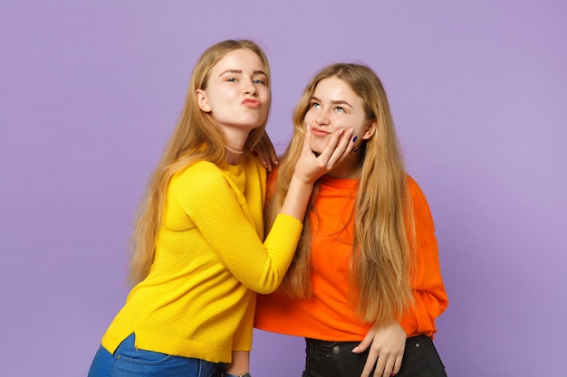 Two cheerful young blonde twins sisters girls in vivid colorful clothes having fun, fooling around isolated on pastel violet blue wall . People family lifestyle concept. 